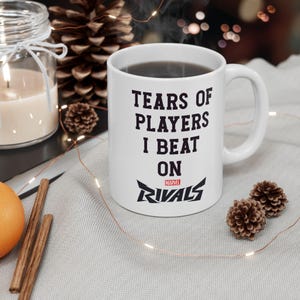 Puede incluir: Taza de cerámica blanca con el texto negro "TEARS OF PLAYERS I BEAT ON RIVALS". La taza tiene un asa curva y está llena de un líquido oscuro. El logotipo de Marvel Rivals está en la parte inferior.