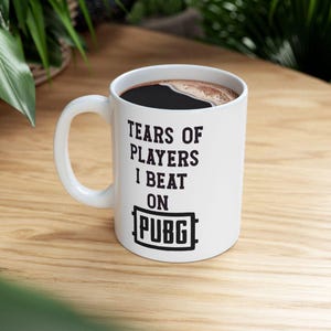 May include: White ceramic coffee mug with the text "TEARS OF PLAYERS I BEAT ON PUBG" in black. The mug is filled with dark coffee and sits on a wooden surface.
