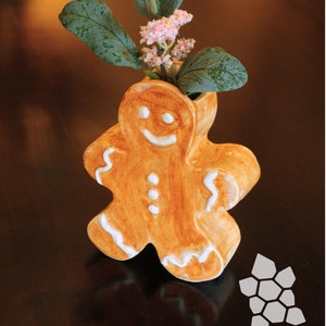 Può includere: Un vaso in ceramica a forma di uomo di pan di zenzero con un viso sorridente e dettagli di glassa bianca, che tiene un mazzo di fiori rosa con foglie verdi.