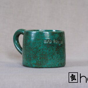 Peut inclure: Une tasse en céramique verte avec un glaçage moucheté et une anse. La tasse porte l'inscription "ars longa" sur le côté.