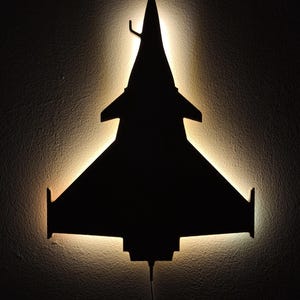 Pode incluir: Silhueta preta de uma luminária de parede em forma de caça em fundo escuro. A luz emite um brilho quente nas bordas, destacando a forma da aeronave. Um cabo de alimentação se estende da parte inferior.