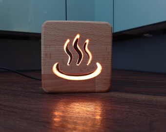 May include: A square wooden lamp with a carved-out design of three curved lines above a curved line, illuminated with a warm glow. The lamp is made of light-colored wood and sits on a wooden surface.