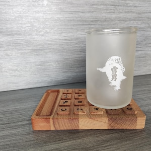 Könnte beinhalten: Ein gefrostetes Glas mit einer weißen Grafik eines Wesens, das einen Hut trägt. Das Glas steht auf einem hölzernen Rechner-Untersetzer mit schwarzen Zahlen und Symbolen.