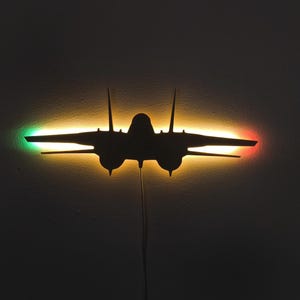 Könnte beinhalten: Schwarze Silhouette eines Kampfflugzeugs mit grünen und roten Lichtern an den Flügeln. Das Flugzeug wird von einem gelb-orangen Licht beleuchtet. Das Bild ist vor dunklem Hintergrund.