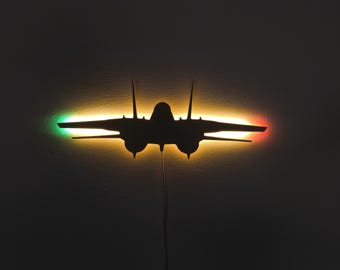 May include: Silhouette of a fighter jet illuminated with green and red lights on the wings. The jet is black, with a yellow and orange glow behind it. The image is set against a dark background.