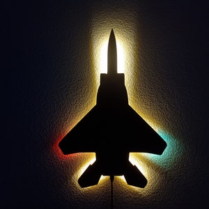 Puede incluir: Una silueta negra de un avión de combate con una luz que brilla a través de las alas, creando un resplandor rojo, amarillo y azul.