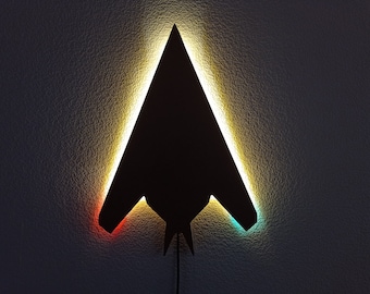 May include: A black silhouette of a stealth fighter jet with a yellow, red, and blue light outline. The light is illuminating the wall behind the silhouette.