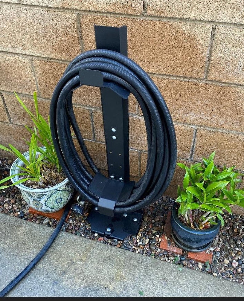 May include: Black metal hose holder mounted to a brick wall with a garden hose wrapped around it. A potted plant is on either side of the holder.
