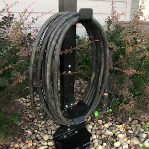 May include: Black metal hose holder with a hose wrapped around it. The holder is standing on a bed of small rocks.