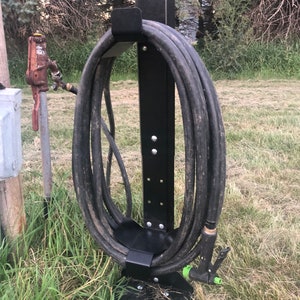 May include: Black metal hose holder with a black hose wrapped around it. The hose holder is standing on a grassy surface.