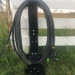 May include: Black metal hose holder with a coiled garden hose attached. The holder is mounted to a white wooden fence and is standing on a bed of green grass.