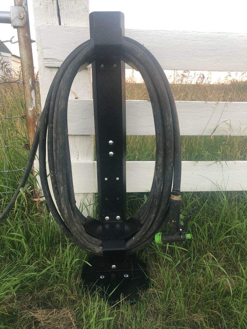 May include: Black metal hose holder with a coiled garden hose attached. The holder is mounted to a white wooden fence and is standing on a bed of green grass.