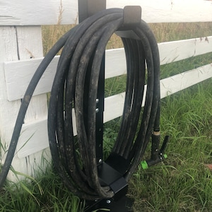 May include: Black metal hose holder mounted to a white fence. The hose is coiled around the holder and the nozzle is hanging down.