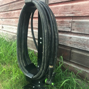 May include: Black metal hose holder with a black hose wrapped around it. The holder is mounted on a wooden surface and is in a grassy area.