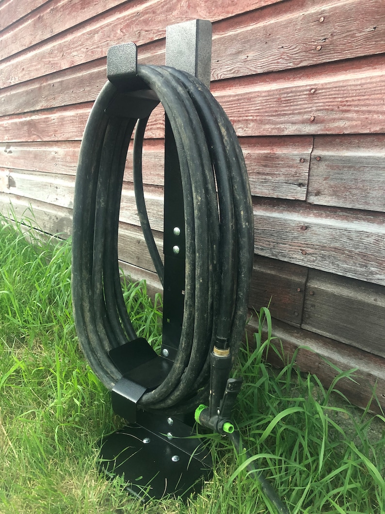May include: Black metal hose holder with a black hose wrapped around it. The holder is mounted on a wooden surface and is in a grassy area.
