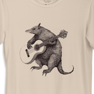 May include: A beige t-shirt featuring a black and white illustration of an armadillo playing a guitar.