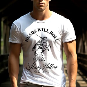 May include: A white t-shirt featuring a black graphic of a person riding a horse. The graphic includes the text "Heads Will Roll" and "Sleepy Hollow New York".