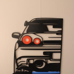Könnte beinhalten: Ein schwarzer Wandbehang mit der stilisierten Frontsilhouette eines Autos. Das Design umfasst zwei markante kreisförmige orangefarbene Scheinwerfer, einen Seitenspiegel und eine dünne horizontale blaue Akzentlinie am unteren Rand, aufgehängt an einer weißen Schnur.