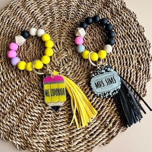 May include: Two beaded wristlet keychains. One features a yellow pencil charm with "MS. CORONADO" text and a yellow tassel. The other has a composition book charm with "MRS. SIMS" text and a black tassel. The beads are in various colors.