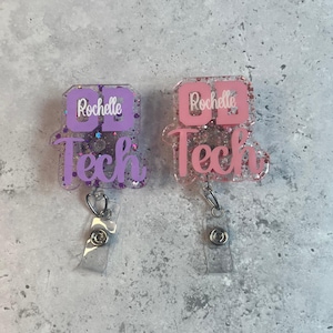 May include: Two glittery badge reels with the text "CD Tech" and the name "Rochelle". One reel is purple and the other is pink. Each reel has a clear plastic strap and a metal clip.
