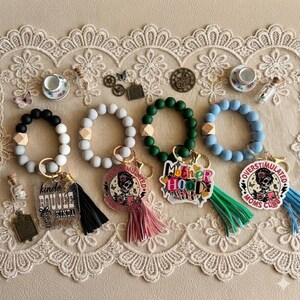 May include: A collection of beaded keychains in various colors, including black, gray, green, and blue. Each keychain features a tassel and a charm with phrases like "kinda boujee kinda broke", "Motherhood", and "Overstimulated Moms Club".