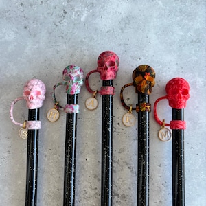 May include: A collection of five black pens, each topped with a decorative skull. The skulls vary in color and pattern, including pink, floral, and orange designs. Each pen has a matching charm with a letter.