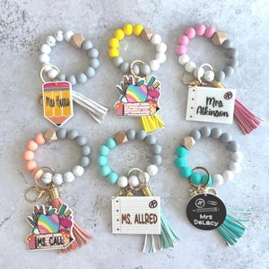 May include: A collection of teacher wristlet keychains. Each keychain features a silicone bead bracelet in various colors, a wooden bead, a tassel, and a charm with a teacher's name or school-themed design.