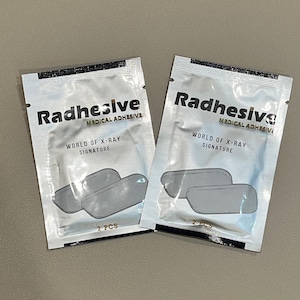 May include: Two white packets of Radhesive medical adhesive, each containing two clear, rectangular adhesive pads. The packets have black text that reads "Radhesive Medical Adhesive" and "World of X-Ray Signature". Each packet contains two adhesive pads and says "2 PCS".