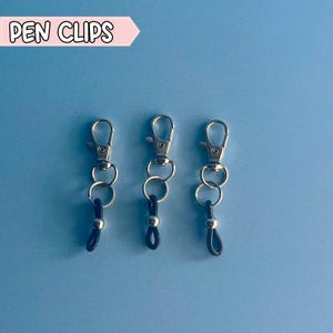 May include: Three silver pen clips with black elastic loops. The clips are attached to the loops with a silver bead.