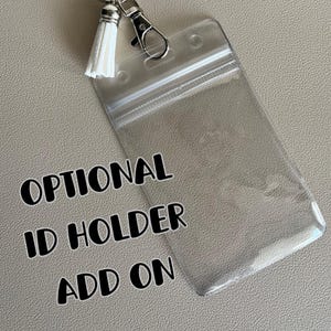 May include: Clear plastic ID holder with a zipper closure. The holder is attached to a silver key chain with a white tassel. The text "OPTIONAL ID HOLDER ADD ON" is printed on the image.