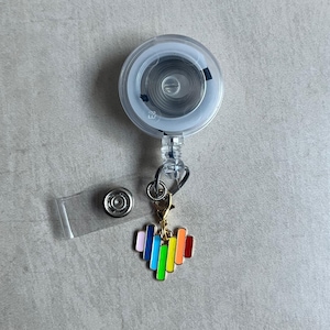 May include: A clear retractable badge reel with a silver metal clip and a rainbow heart charm. The charm features vertical bars in various colors, including pink, blue, green, yellow, and red. A clear plastic snap is also included.