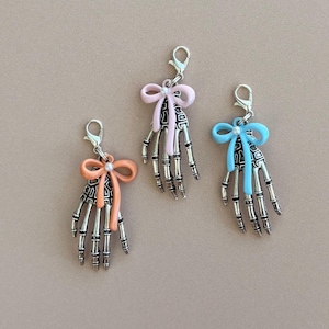May include: Three skeleton hand charms with lobster clasps. Each charm features a silver-toned skeletal hand design with a colorful bow and a small pearl accent. The bows are in shades of peach, pink, and blue.