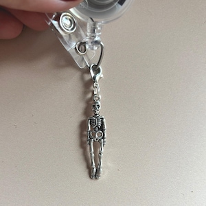 May include: A silver-colored skeleton charm attached to a clear plastic retractable badge reel. The skeleton has detailed rib cage and leg bone features. The reel has a metal clip and a small metal clasp connecting the charm.
