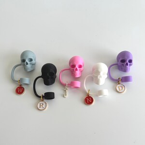 May include: A collection of colorful skull-shaped bottle stoppers. Each stopper has a different color skull, including blue, black, pink, white, and purple. Each stopper has a loop and a charm with a letter.