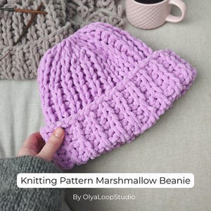May include: A chunky knit beanie in a light lavender color with a thick folded brim. The beanie is made from a soft, plush yarn with a visible looped texture. The text Knitting Pattern Marshmallow Beanie By OlyaLoopStudio is visible.