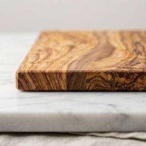 May include: A rectangular wooden cutting board with a natural wood grain pattern. The board is made of olive wood and has a light brown color with darker brown streaks. The board is resting on a white marble surface.