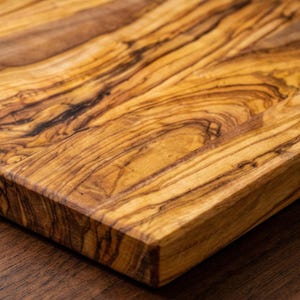 Puede incluir: Primer plano de una tabla de cortar de madera con un patrón de vetas naturales. La tabla tiene forma rectangular con un borde biselado. La madera es de color marrón claro cálido con vetas y nudos marrones más oscuros.