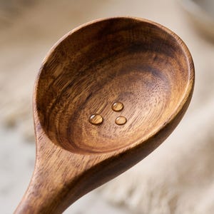 Puede incluir: Una cuchara de madera profunda de forma ovalada con un rico patr&oacute;n de vetas marrones. El interior del cuenco de la cuchara presenta tres peque&ntilde;as gotas de agua transparentes sobre la superficie lisa y pulida, resaltando la textura natural.