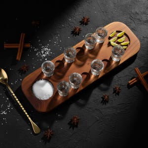 Puede incluir: Una bandeja de madera con ocho vasos de chupito llenos de líquido transparente. La bandeja también contiene un pequeño cuenco de sal, gajos de lima y una cuchara dorada. Palitos de canela y anís estrellado esparcidos.