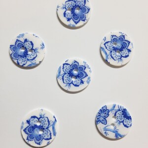 May include: Six round, white buttons with a blue floral design. Each button features a detailed blue flower in the center, surrounded by smaller floral accents. The buttons are arranged on a white surface.