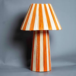 Può includere: Una lampada da tavolo decorativa con base conica e paralume. La lampada presenta un motivo a strisce verticali arancioni e bianche. La lampada è un apparecchio di illuminazione decorativo.