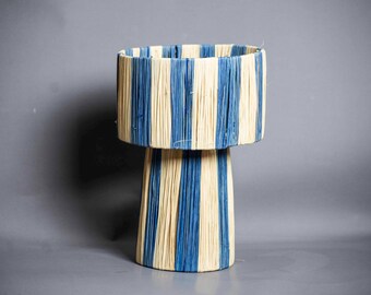 May include: A decorative lamp with a cylindrical base and a wider, drum-shaped shade. The lamp features vertical stripes of blue and cream-colored material, creating a textured, striped pattern. The lamp is a unique home decor item.