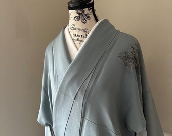 Può includere: Un kimono azzurro con un motivo floreale sulla manica è esposto su un manichino. Il kimono ha una chiusura a portafoglio e una fodera bianca. Il manichino ha una testa nera e un busto color crema.