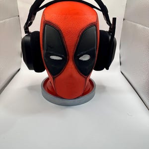 May include: A red and black Deadpool-themed headphone stand. The stand is shaped like Deadpool's head, with black eye shapes and a textured surface. Black headphones rest on the stand, which sits on a gray circular base.