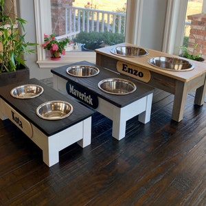 Peut inclure: Trois bols sur&eacute;lev&eacute;s pour chien avec des noms personnalis&eacute;s "Kota", "Maverick" et "Enzo". Les bols sont en acier inoxydable et sont pos&eacute;s sur des supports en bois avec une finition noire et blanche.
