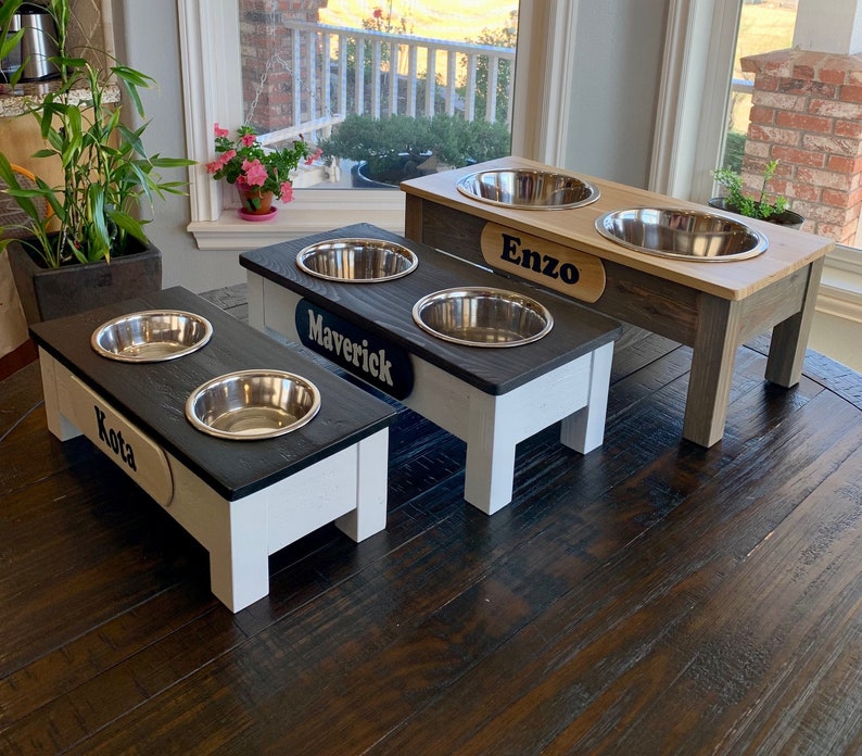 Peut inclure: Trois bols sur&eacute;lev&eacute;s pour chien avec des noms personnalis&eacute;s "Kota", "Maverick" et "Enzo". Les bols sont en acier inoxydable et sont pos&eacute;s sur des supports en bois avec une finition noire et blanche.