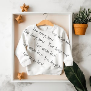 May include: White long-sleeve shirt with the text "Your design here!" repeated on the front. The shirt is hanging on a wooden hanger in a white frame.