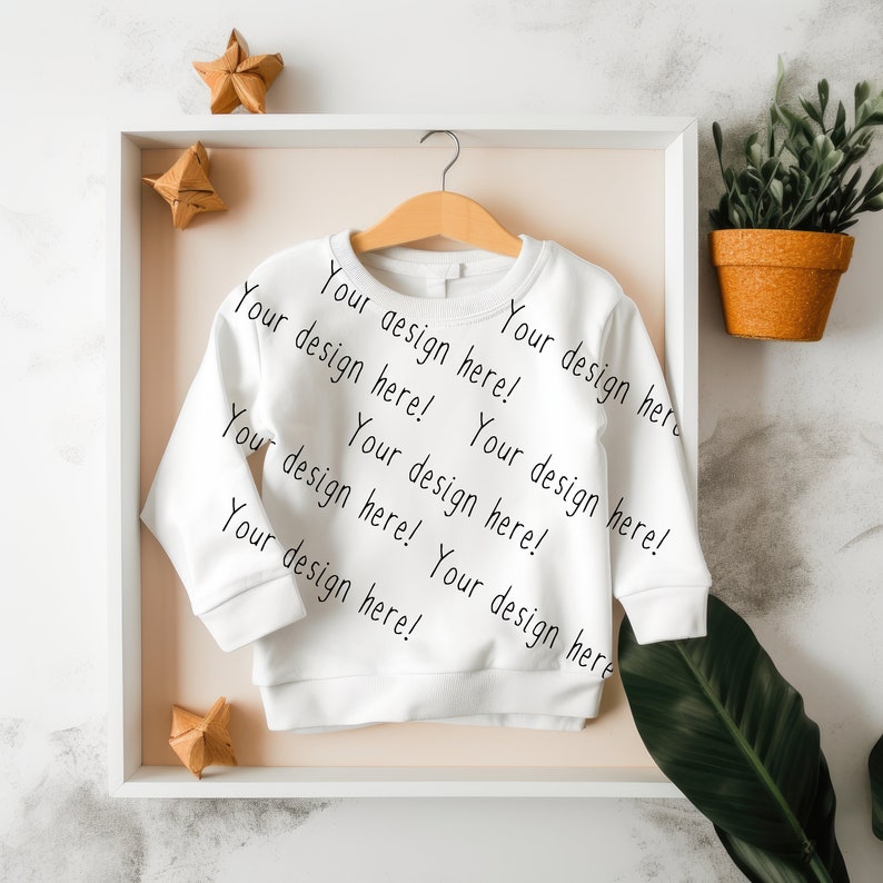May include: White long-sleeve shirt with the text "Your design here!" repeated on the front. The shirt is hanging on a wooden hanger in a white frame.
