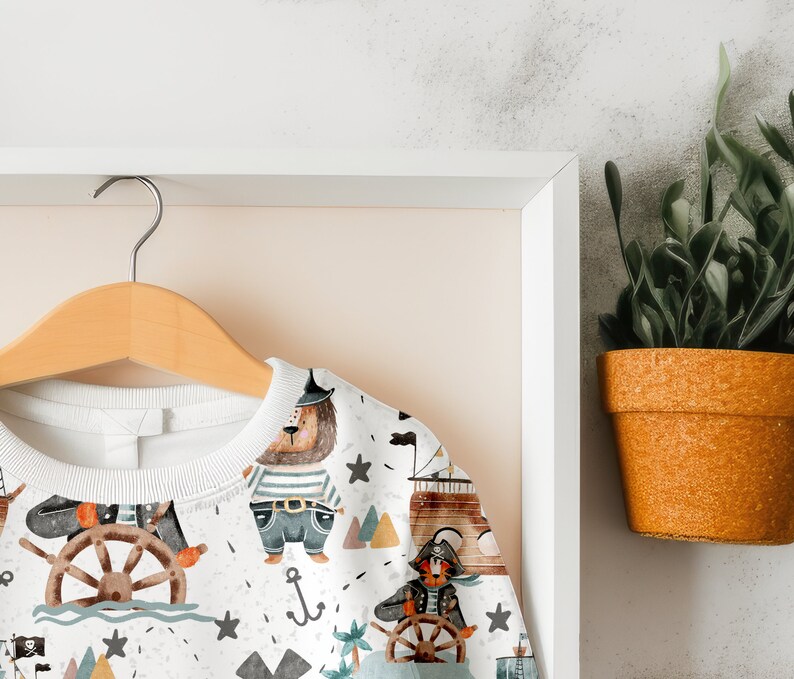 May include: White sweatshirt with a colorful pattern of animals, ships, and anchors. The pattern includes a bear wearing a pirate hat, a tiger wearing an eye patch, and a ship with a sail.