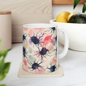Può includere: Tazza in ceramica bianca con un design ad acquerello di neuroni rosa, rossi e blu su uno sfondo beige.